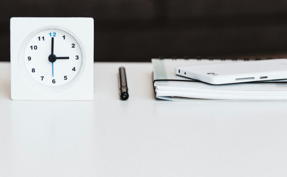 Schreibtisch mit Uhr und Arbeitsmaterialien wie Füller, Handy und Arbeitsmappe