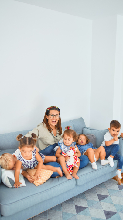 Zu sehen ist eine lächelnde junge Frau, die mit fünf kleinen Kindern auf einer blauen Kautsch sitzt.