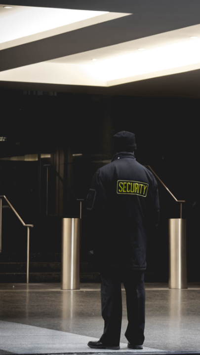 Ein Mann steht mit dem Rücken zur Kamera in einem Foyer, auf dem Rücken seiner dunklen Jacke steht in Schulterhöhe in gelber Schrift "SECURITY".