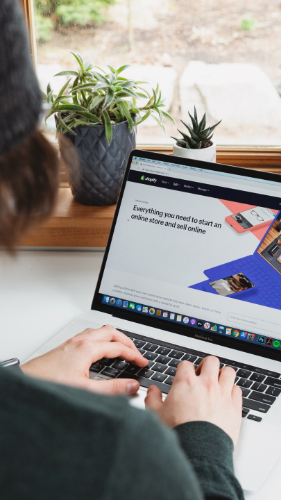 Aus der Schulterperspektive sieht man Hände, die etwas auf der Tastatur des Laptops eingeben. Auf dem Bildschirm des Laptops ist die Internetseite "Shopify" zu sehen.